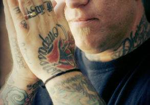 A bald man with tattoos on his arms and hands, inspired by the Spiritual teachings of Noah Levine, holding his hands together near his face, looking at the camera.