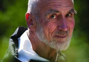 Elderly man with a white beard and balding head, wearing a black and white hooded garment, embodying gratitude practice as he looks over his shoulder in a sunlit natural setting.