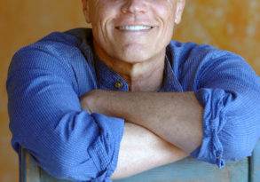 Middle-aged man with glasses smiling at the camera, sitting with crossed arms on a blue chair against an orange textured background featuring posters about climate solutions.