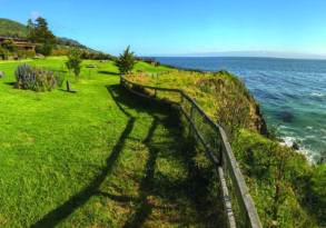 Panoramic view of a lush green cliffside with a wooden fence leading into the mystic ocean under a clear blue sky.