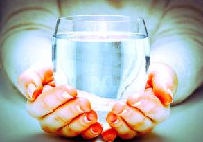 Two hands with orange fingernails holding a clear glass of life-giving water, with a soft-focus background.