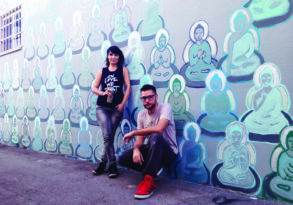 Two people posing in front of a colorful spiritual graffiti with multiple buddha figures in various poses.