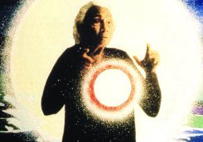 A magician in a black robe gestures dramatically in front of a large, glowing circular screen that emits sparkling light effects, uttering his last words.