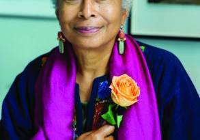 Portrait of an elderly woman with gray hair, smiling gently at the camera, holding an orange rose, wearing a purple shawl and "Womanist" earrings.