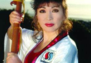 A woman with dark hair wearing a white and red martial arts uniform holds a wooden staff, posing outdoors with a scenic background. This is Tae Yun Kim.