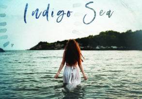 A woman with long red hair wades into the ocean, facing away and wearing a white dress, with the text "Indigo Sea Ajeet Kaur" overlaid in artistic font. The