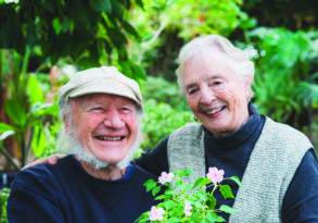 Elderly couple smiling and holding a potted flowering plant together in a lush garden, reminiscent of a scene from a family TV show.