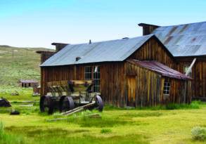 Old wooden farm buildings with a rusty wagon in a grassy field, depicting a deserted rural area under a clear sky.