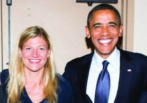 Two smiling people, one woman and one man in a suit, meet for a photo indoors.