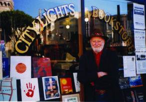 Elderly man with a beard and hat standing in front of city lights bookstore, smiling, with arms crossed. Book displays and posters visible through the window, including one featuring a new release titled