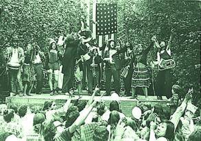 A group of performers at the Oregon Country Fair on a woodland stage engaging with a lively audience, featuring musicians and dancers in casual attire celebrating the 50th Anniversary.