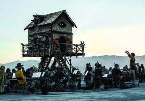 A rustic wooden house on stilts at the Burning Man festival, surrounded by people and musicians in the desert at sunset.