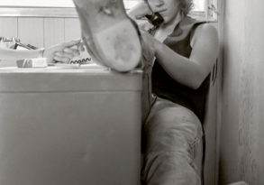 A young person lounges casually at the Woodstock Music Festival, one leg propped up on a counter, talking on a phone in a cluttered room, displaying a worn-out sole of a sne