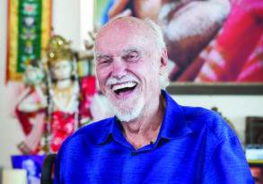 Elderly man in a blue shirt laughing joyfully during his last conversation while sitting in a wheelchair, with a colorful painting and a religious statue in the background.