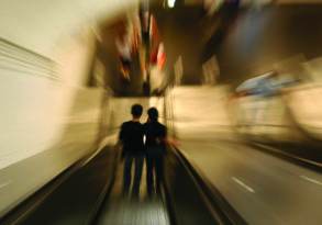 A blurred image of two people holding hands, embodying love as they stand on a moving escalator in a tunnel with bright lights and motion blur effects.