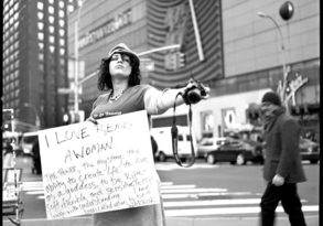 A person in a city street holding a sign that says "i love being a woman" and a camera, with pedestrians and tall buildings in the background, radiating female strength.