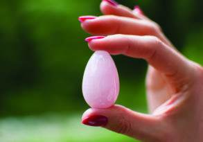 Close-up of a hand with red nail polish holding a small, smooth pink yoni egg against a blurred green background.