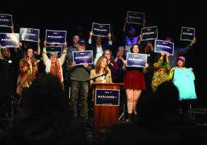 A woman speaks at a podium with a group of people holding signs with the name "Marianne Williamson" in a crowded auditorium.