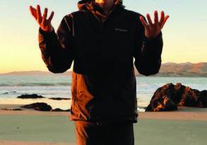 A man balancing three rocks stands on a beach at sunset, with mountains visible in the background.