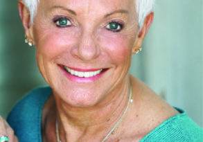 An elderly woman with short white hair and sparkling eyes, smiling broadly, *participates in a channeled conversation*, wearing a turquoise top and silver jewelry.