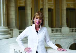 A woman in a white blazer, embodying resilience, smiling at the camera while standing next to a marble column in an elegant hallway.