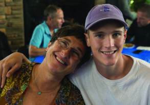 A smiling middle-aged woman with glasses, known for her profession as an editor, embracing a young man wearing a blue cap, both sitting at a table in a casual setting.