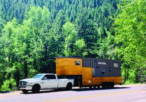 A white pickup truck towing a large wooden tiny home on wheels parked on a road next to a dense green forest.