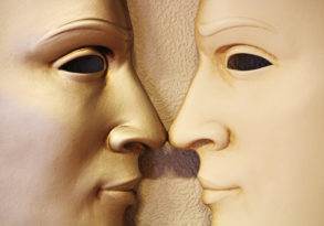Two white face masks closely positioned as if touching noses against a textured cream background, symbolizing the interplay of epigenetics in health.