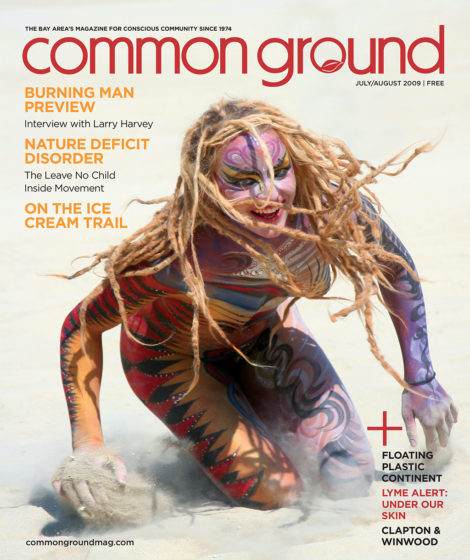 A person with dreadlocks, wearing tribal body paint, is crawling on sandy ground, featured on the July/August 2009 cover of Common Ground magazine.