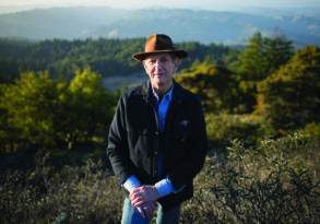 A man in a hat and coat stands in a forested area with rolling hills in the background during sunset, embodying a sense of zen.