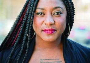 Portrait of a woman with braided hair and a visible tattoo on her chest, embodying the spirit of a Freedom Dreamer, wearing a blue top and black cardigan, standing outdoors.