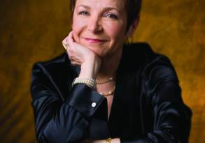Caroline Myss, a woman with short red hair, smiles at the camera, resting her chin on her hand against a golden background. She wears a black jacket and pearl earrings.
