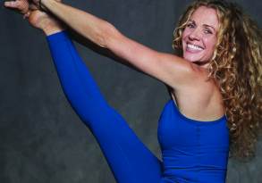 Woman in blue athletic wear performing a seated yoga pose, smiling at the camera, in front of a gray backdrop, demonstrating Seane Corn's yoga teachings.
