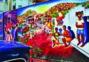 Colorful street mural depicting a vibrant community scene with buildings, people engaging in daily activities, and a large mythical figure on the left, symbolizing spiritual engagement.