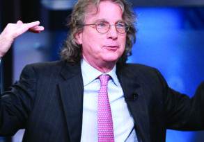 Middle-aged man with long curly hair wearing a suit and glasses, gesturing with his hands while speaking about parenting, against a blurred background.