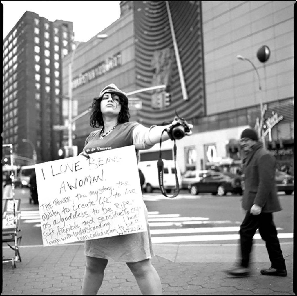 woman with camera and poster I Love Being a Woman - Layla Love