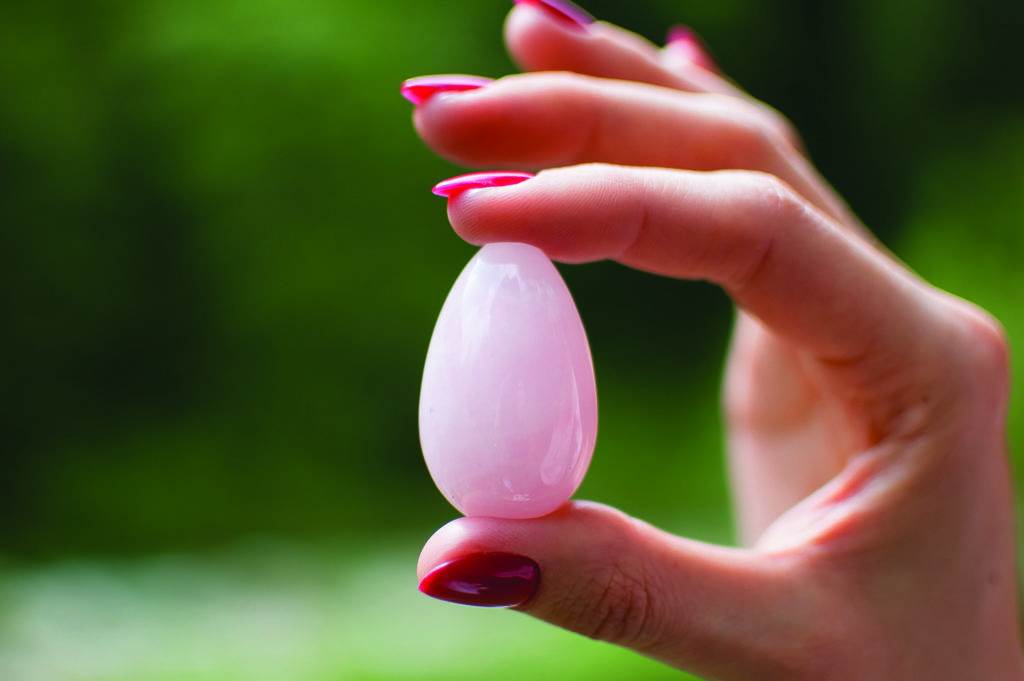 Close up photo of female hand holding a yoni egg. Rose quartz crystal egg on river background. Womens health concept.