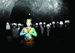 A colorized woman in traditional attire illuminated in a cave surrounded by people in hard hats and white robes uttering their last words, all in grayscale.