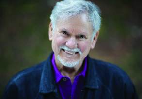 An elderly man with white hair and a beard, smiling warmly at the camera, wearing a purple shirt and a black jacket, with blurred greenery in the background. The serene expression and thoughtful demeanor resemble