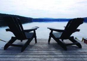 Two adirondack chairs facing a tranquil lake, set on a wooden dock with a forested hill in the background, offering perfect relaxation.